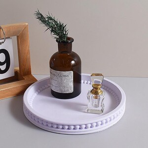 May include: A round, light purple decorative tray with a beaded edge holds a brown glass bottle with a sprig of greenery and a small, clear perfume bottle with a gold cap. A wooden calendar frame is partially visible.