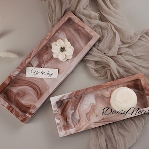 Op de afbeelding: Twee rechthoekige trays met een gemarmerd, roodbruin en wit ontwerp. Eén tray heeft een witte bloem en een kaart met de tekst "Yesterday". De andere tray heeft een wit, rond object. De trays staan op een lichtgrijze ondergrond.