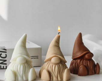 Puede incluir: Tres velas con forma de gnomo en crema, beige y marrón. La vela central tiene una mecha encendida. Los gnomos tienen largas barbas y sombreros puntiagudos. Las velas están sobre una superficie blanca.