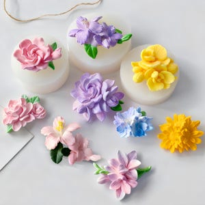 Op de afbeelding: Een verzameling kleurrijke, kunstmatige bloemdecoraties in verschillende vormen en maten. De bloemen zijn in tinten roze, paars, geel en blauw, met groene bladeren. Sommige staan op witte, ronde bases.