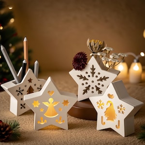 Puede incluir: Una colección de adornos blancos en forma de estrella. Algunas estrellas tienen diseños recortados de copos de nieve, ángeles y otras formas festivas. Lápices y flores secas se colocan en algunas de las estrellas. Los artículos están iluminados con un brillo cálido.