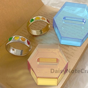 Puede incluir: Dos anillos con diseños de esmalte en forma de corazón colorido y dos expositores hexagonales de acrílico. Un anillo es plateado, el otro dorado. Los soportes son azules y naranjas translúcidos. Los artículos están sobre una superficie marrón.