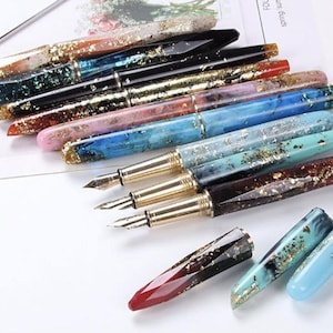 Può includere: Una collezione di penne stilografiche con pennini color oro e fusti decorativi. I fusti presentano una varietà di colori, tra cui blu, rosa e nero, con accenti di foglia d'oro. Le penne sono disposte su una superficie bianca.