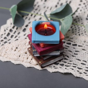 May include: A stack of miniature book-shaped candles in various colors, topped with a lit blue candle. The candle flame glows brightly against the dark red wax. The books are in shades of brown, red, and orange.