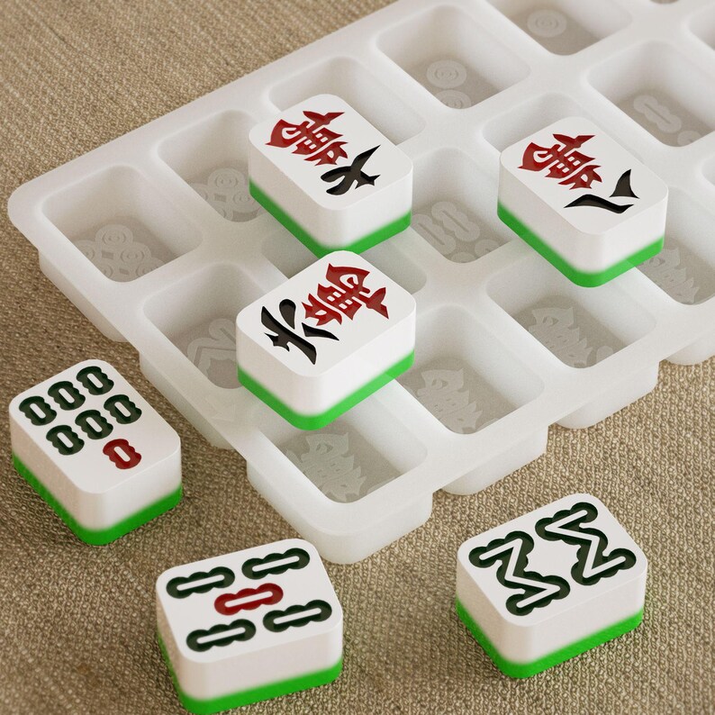 May include: A collection of white Mahjong tiles with green edges, featuring red and black Chinese characters and symbols. The tiles are arranged in a clear, grid-like tray, suggesting a game setup. The background is a neutral, textured surface.