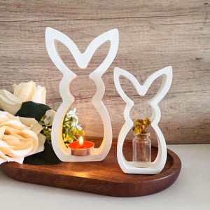 May include: Two white bunny-shaped candle holders on a wooden tray. One holds a lit orange candle, the other a small glass vase with dried yellow flowers. Decorative Easter or spring home decor.
