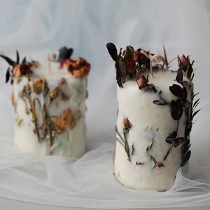 May include: Two white pillar candles decorated with dried flowers and foliage. The candles have a textured appearance with the botanical elements embedded in the wax. The flowers are in shades of orange, red, and brown.