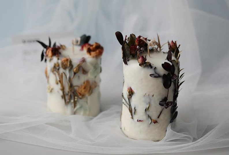 May include: Two white pillar candles decorated with dried flowers and foliage. The candles have a textured appearance with the botanical elements embedded in the wax. The flowers are in shades of orange, red, and brown.