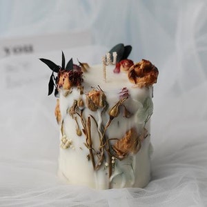 May include: A white, cylindrical candle decorated with dried flowers and foliage. The candle features a variety of textures and colors, including brown, red, and green, creating a natural and rustic aesthetic. The candle is a decorative item.