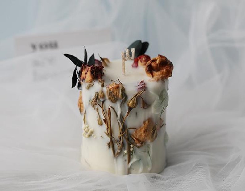 May include: A white, cylindrical candle decorated with dried flowers and foliage. The candle features a variety of textures and colors, including brown, red, and green, creating a natural and rustic aesthetic. The candle is a decorative item.