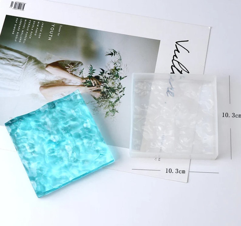 May include: A square, clear resin mold with a floral design, next to a finished square resin piece in shades of blue. The mold and finished piece are approximately 4 inches in size. A magazine with the word "YOUTH" is in the background.
