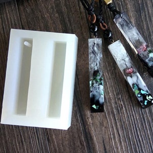 Peut inclure: Un moule en silicone rectangulaire blanc avec deux cavités verticales, à côté de trois pendentifs rectangulaires en résine. Les pendentifs sont transparents avec des motifs incrustés, dont des fleurs et des motifs abstraits, suspendus à des cordons noirs.
