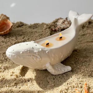 Puede incluir: Un portavelas blanco con forma de ballena con dos velas encendidas. La ballena de cerámica tiene una superficie texturizada y descansa sobre arena. Conchas marinas están esparcidas alrededor del portavelas, creando un tema playero.