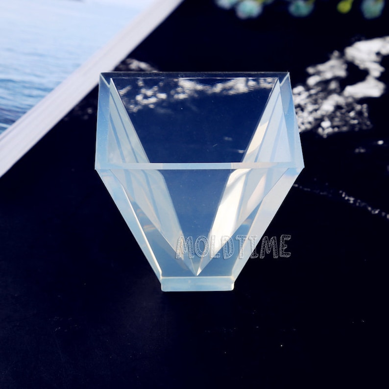 May include: A clear, geometric mould with a diamond-like shape. The mould has a wide opening and angled sides, with the text "MOLDTIME" visible on the front. The mould is made of a translucent material.