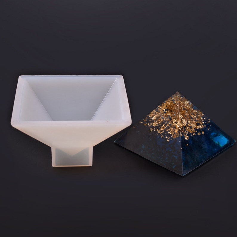 May include: A white silicone pyramid mold next to a finished pyramid. The finished pyramid is dark blue and gold, with gold leaf flakes on the top. The mold is square with a pyramid shape.