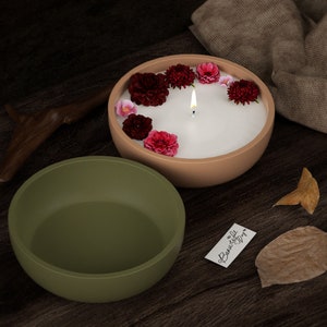 May include: A burning candle in a beige bowl, decorated with red and pink flowers. An olive green bowl is next to it. A small tag with "Beautiful Day" is visible. The items are on a dark wooden surface.