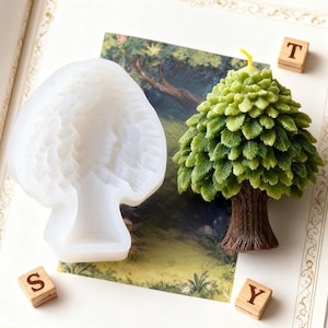 May include: A tree-shaped candle with a brown trunk and green leaves, next to a white silicone mold in the shape of a woman's face. Wooden letter blocks spell out "STY". The candle has a yellow wick.
