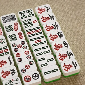 May include: A collection of white Mahjong tiles with black, red, and green accents. The tiles feature various symbols and Chinese characters. The game pieces are arranged on a neutral-colored surface.