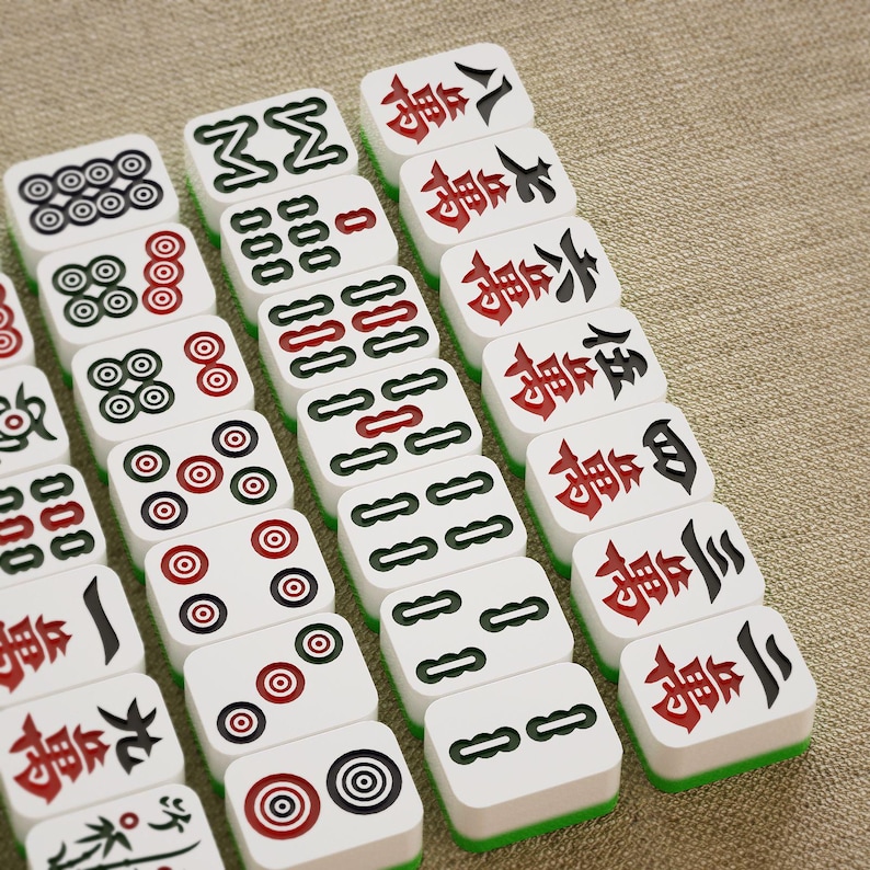 May include: A collection of white Mahjong tiles with black, red, and green accents. The tiles feature various symbols and Chinese characters. The game pieces are arranged on a neutral-colored surface.