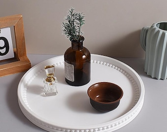 Può includere: Un vassoio decorativo rotondo bianco con bordo perlato contiene una piccola ciotola marrone, un flacone di profumo trasparente e un vaso di vetro marrone con verde. Sono presenti anche un calendario con cornice in legno e un vaso di ceramica azzurra.