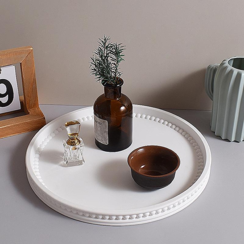 May include: A white, round decorative tray with a beaded edge holds a small brown bowl, a clear perfume bottle, and a brown glass vase with greenery. A wooden framed calendar and a light blue ceramic vase are also present.