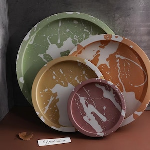 May include: A collection of four round decorative trays in different sizes and colours. Each tray has a white splatter paint design on a green, gold, orange, and mauve background. A small card with the word "Yesterday" is visible.
