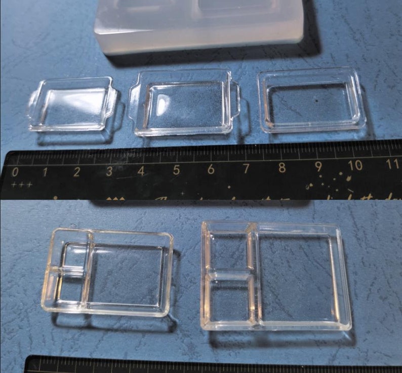 May include: Clear silicone moulds for crafting. The image shows several small, clear trays with varying designs, including some with handles and divided compartments. A ruler is included for scale, showing the moulds are small.