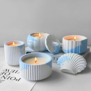 Puede incluir: Una colección de velas encendidas en recipientes decorativos. Las velas están en recipientes veteados blancos y azules, incluyendo una vela con forma de calabaza y una vela con forma de concha. Las velas están sobre una superficie blanca.