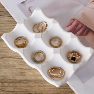 May include: A white ring holder with six compartments, containing several gold and silver rings. The rings vary in design, including bands, a ring with a black square stone, and a ring with pearls. The holder is on a white surface.