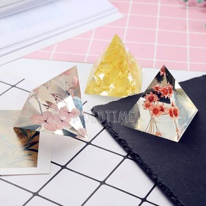 May include: Three clear resin pyramids, each with dried flowers inside. One has pink flowers, another yellow, and the third red. The pyramids are displayed on a patterned surface.