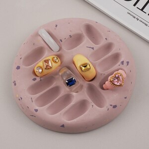 Può includere: Una tavolozza per nail art rosa a forma ovale con più rientranze, contenente vari design di unghie gioiello e un anello. Le unghie presentano basi trasparenti, gialle e rosa con accenti dorati e gioiello. Sono presenti anche un'unghia bianca e un anello rosa a forma di cuore.