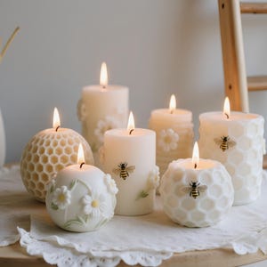 May include: A collection of lit, ivory-colored candles with various designs. Some candles feature honeycomb patterns, floral accents, and bee decals. The candles are arranged on a white lace cloth, creating a warm and inviting ambiance.