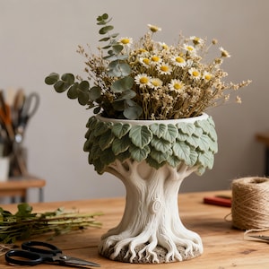 Puede incluir: Una maceta blanca con forma de árbol con un borde verde en forma de hoja, llena de flores secas y vegetación. La maceta tiene un diseño de corteza texturizada y raíces visibles en la base. El arreglo se encuentra sobre una superficie de madera, lo que sugiere un tema rústico o inspirado en la naturaleza.