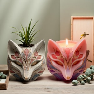 May include: Two fox-shaped decorative items. One is a planter with a plant inside, the other is a candle. Both are in the shape of a fox head with intricate, colorful floral designs. The candle is lit, and the planter is unlit.