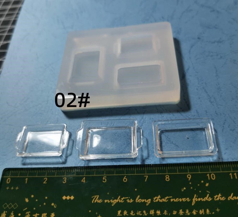 May include: A clear silicone mould with three rectangular cavities and three small, clear rectangular trays. The mould has the text "02#" printed on it. A ruler is at the bottom of the image.