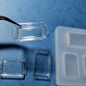 May include: Clear, rectangular miniature baking dishes with handles, one held by tweezers. The image also shows a silicone mould with matching rectangular cavities. The dishes are likely for crafting or miniature projects.