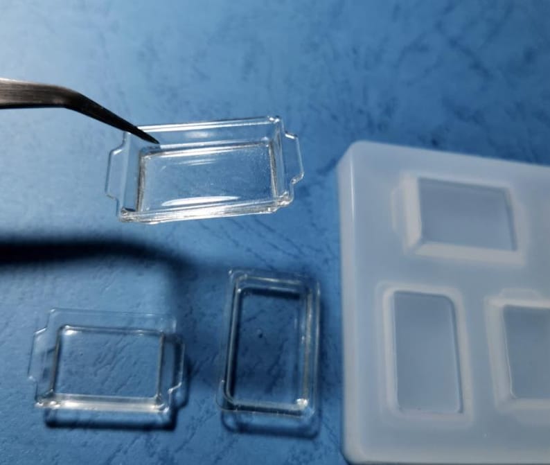 May include: Clear, rectangular miniature baking dishes with handles, one held by tweezers. The image also shows a silicone mould with matching rectangular cavities. The dishes are likely for crafting or miniature projects.