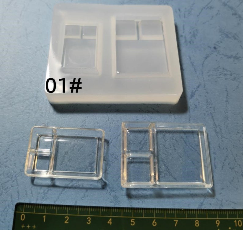 May include: A clear silicone mould with multiple rectangular and square cavities, alongside two cast resin pieces. The mould has the number "01#" printed on it. A ruler is at the bottom, showing measurements in centimetres.