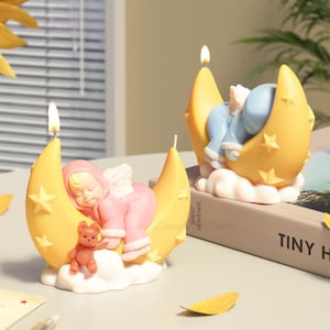 Puede incluir: Dos velas decorativas encendidas con forma de bebé durmiendo en una luna creciente. Una vela presenta un bebé rosa con un osito de peluche, la otra un bebé azul. Ambas están sobre una base de nube con estrellas amarillas.