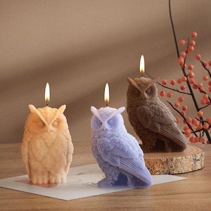 May include: Three owl-shaped candles in shades of beige, lavender, and brown, each with a lit wick. The candles are intricately detailed, showcasing the texture of feathers. One candle is on a wooden slice, and the others are on a white surface.