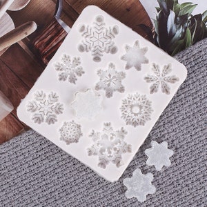 Può includere: Uno stampo in silicone bianco con vari disegni di fiocchi di neve. Lo stampo contiene nove diverse forme di fiocchi di neve, con due fiocchi di neve sciolti accanto. I fiocchi di neve hanno dettagli intricati, adatti per lavori creativi o pasticceria.