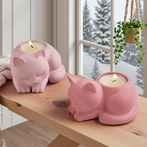 Könnte beinhalten: Zwei rosa katzenförmige Kerzenhalter mit brennenden Kerzen. Die Katzenfiguren befinden sich in Schlafposition, die Augen geschlossen. Die Kerzenhalter stehen auf einer Holzbank in der Nähe eines Fensters mit einer verschneiten Szene.