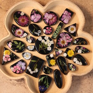 May include: A selection of resin pendants and charms, each featuring pressed flowers in diverse shapes like ovals and triangles. The flowers are in pink, yellow, and white, set against a black backdrop. The items are presented on a light brown, flower-shaped tray.