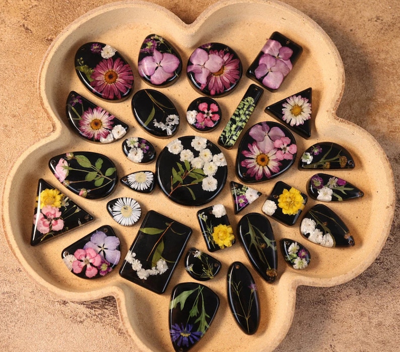 May include: A selection of resin pendants and charms, each featuring pressed flowers in diverse shapes like ovals and triangles. The flowers are in pink, yellow, and white, set against a black backdrop. The items are presented on a light brown, flower-shaped tray.