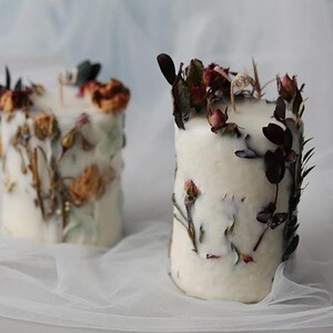 May include: Two white pillar candles decorated with dried flowers and foliage. The candles have a cylindrical shape and are adorned with various dried flowers in shades of red, orange, and green. The candles are placed on a white, sheer fabric.