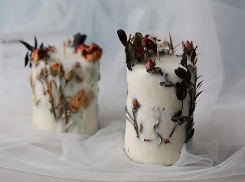 May include: Two white pillar candles decorated with dried flowers and foliage. The candles have a cylindrical shape and are adorned with various dried flowers in shades of red, orange, and green. The candles are placed on a white, sheer fabric.