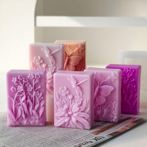 May include: A collection of rectangular soaps in various shades of pink and purple. Each soap bar features a detailed floral and insect relief design, including butterflies, dragonflies, and flowers. The soaps are arranged on a newspaper.