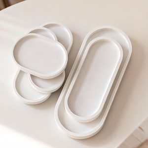 May include: A set of white, oval serving trays in different sizes. The trays are stacked and arranged on a white surface. The design is minimalist and modern, suitable for home decor. The trays are made of a solid, opaque material. The measurements are in centimeters.