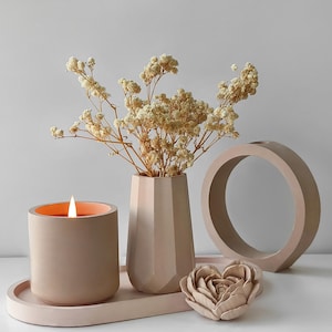 Pode incluir: Uma composição de decoração para casa em tons neutros. Inclui uma vela acesa, um vaso geométrico com flores secas, um anel decorativo e um adorno em forma de rosa. A vela está numa bandeja, em tons de bege e pêssego.