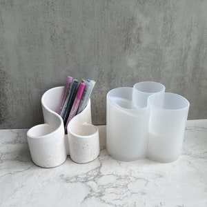 May include: Two white pen holders. One has a unique, curved design with three compartments and contains several pens. The other is a translucent white with three compartments. Both are on a marble surface.